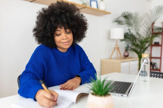 A woman working from home in a modern office, using a laptop and writing in a notebook.