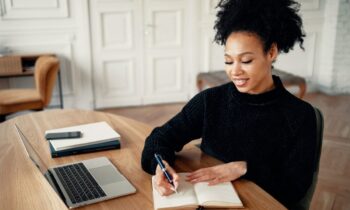 A woman working from her home office desk, using a laptop and writing in a notebook for blog post make money writing short stories