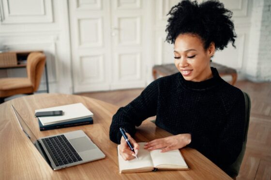 A woman working from her home office desk, using a laptop and writing in a notebook for blog post make money writing short stories