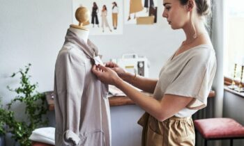A fashion designer working on altering a top on a mannequin in her office, with a sewing machine and fashion illustrations in the background for blog post make money in fashion