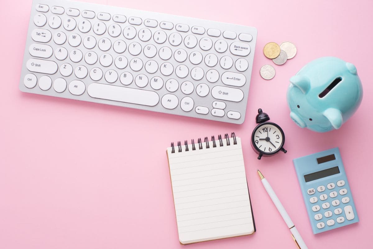 A work from home office desk with a keyboard, notebook, coins, a clock, a calculator, and a piggy bank.