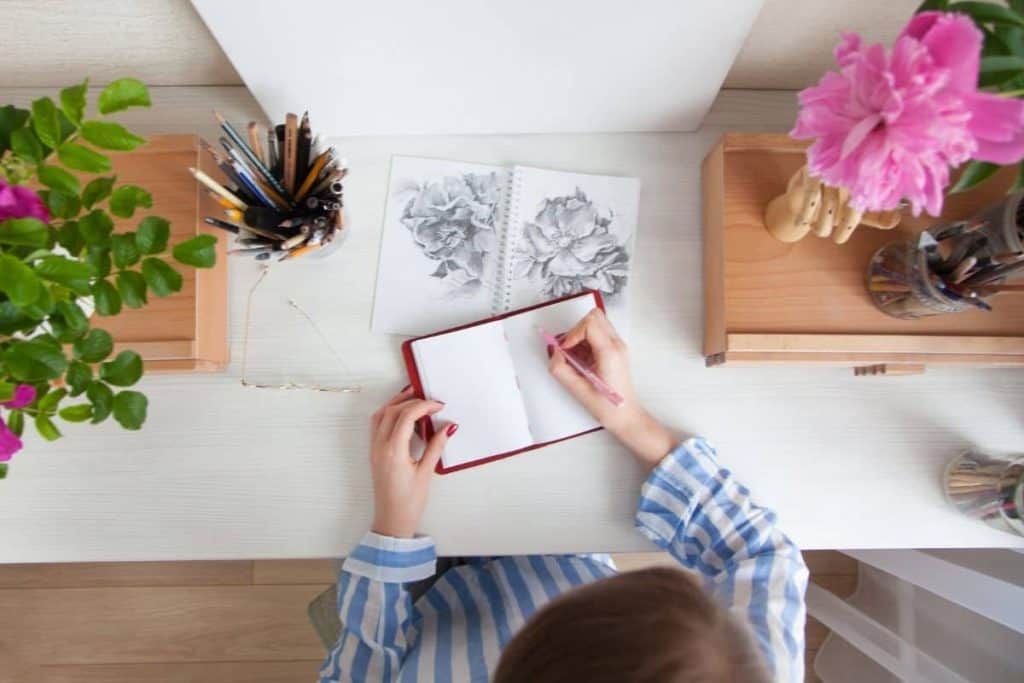 Overhead view of an artist, drawing flowers in a sketchbook at her home office desk.