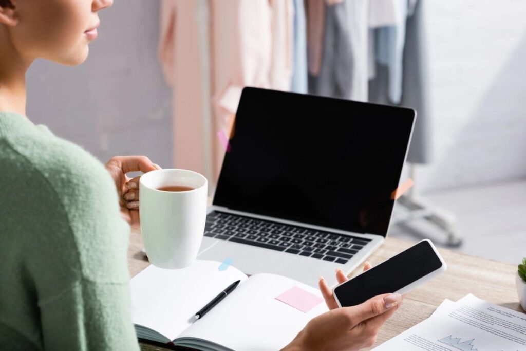 A woman working remotely in her home office, using a laptop and holding a cell phone.