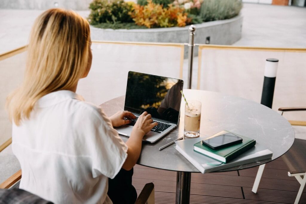 A woman working outdoors at a table, using a laptop and drinking coffee.