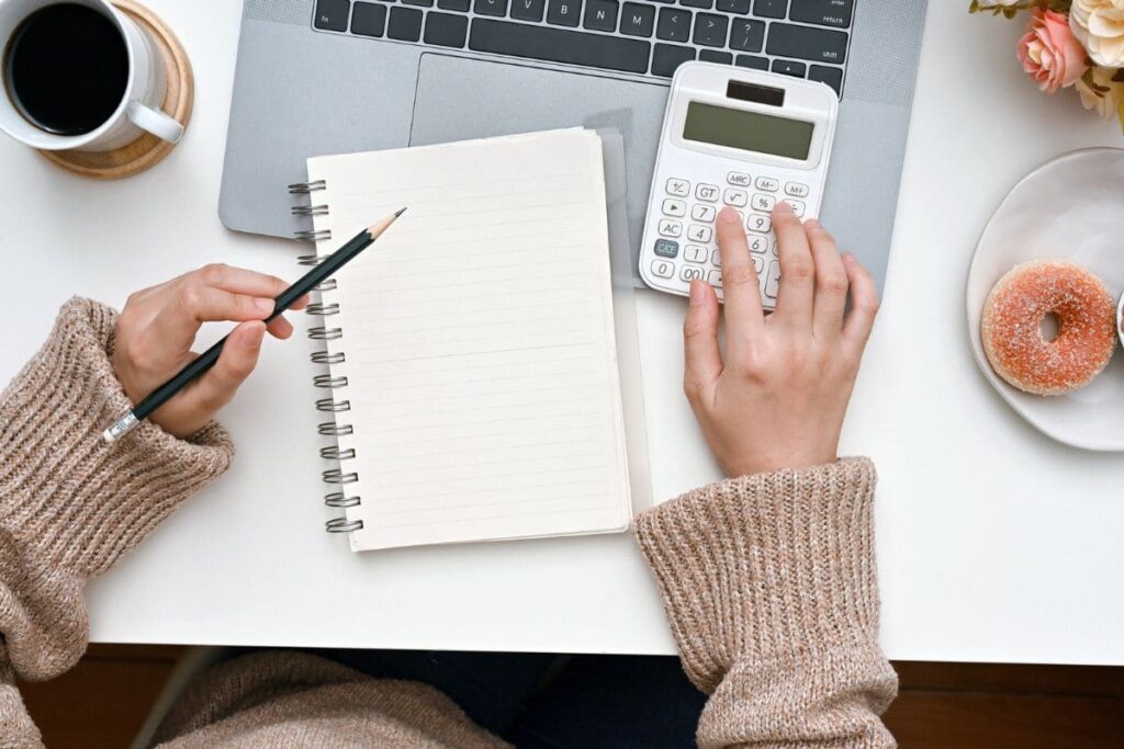 A woman sitting at her home office desk, using a calculator and writing in a notebook.