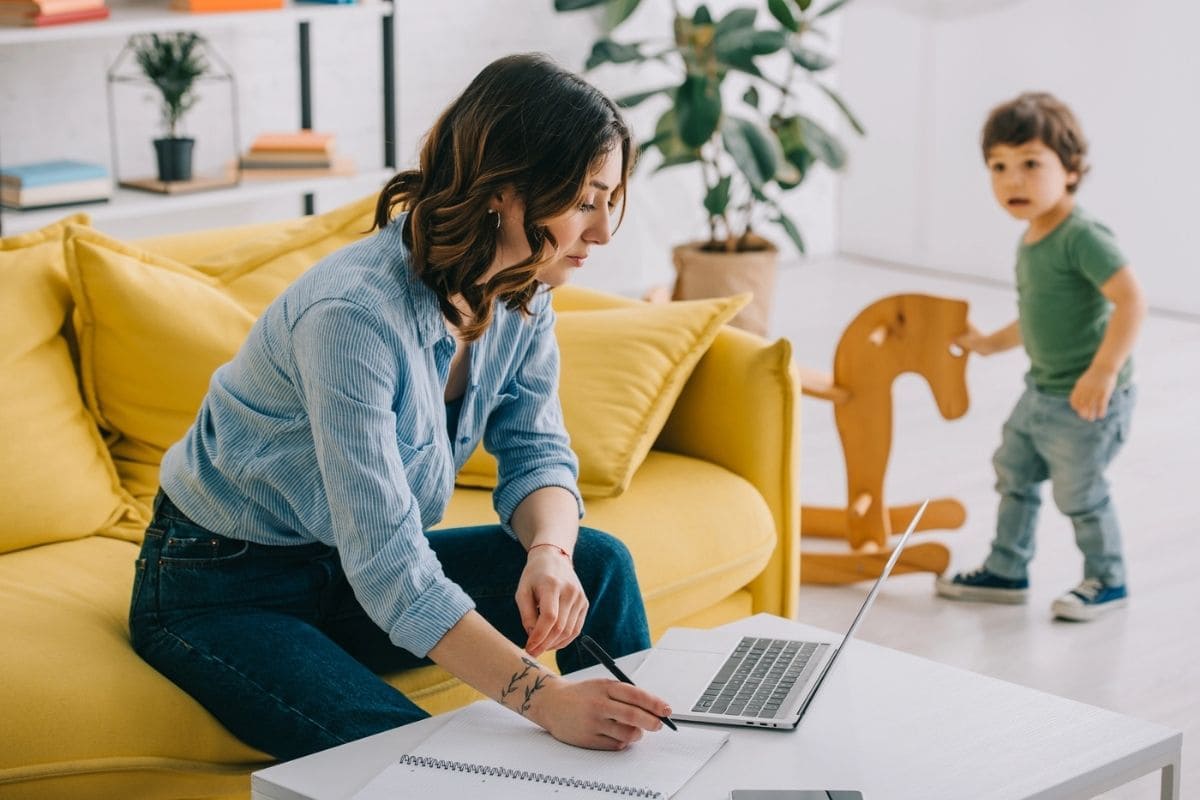 A woman working from home, sitting on a coach and using a laptop while her child plays in the background for blog post how to restart your career after a break