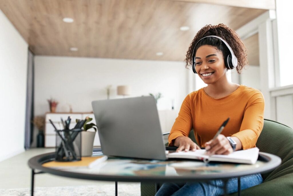 A freelancer working remotely from her kitchen table, using a laptop, wearing headphones, and writing in a notebook.