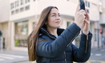 A woman standing outside in the city, using a smartphone to take a photo.