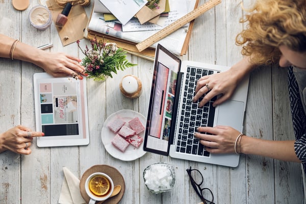 Two women, one working on a tablet, the other working on a laptop sipping tea and eating cookies