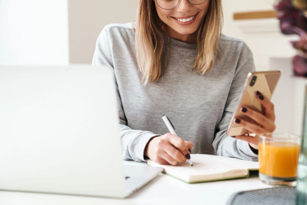 A woman sitting at a desk in her home office, using a smartphone and writing in a notebook.