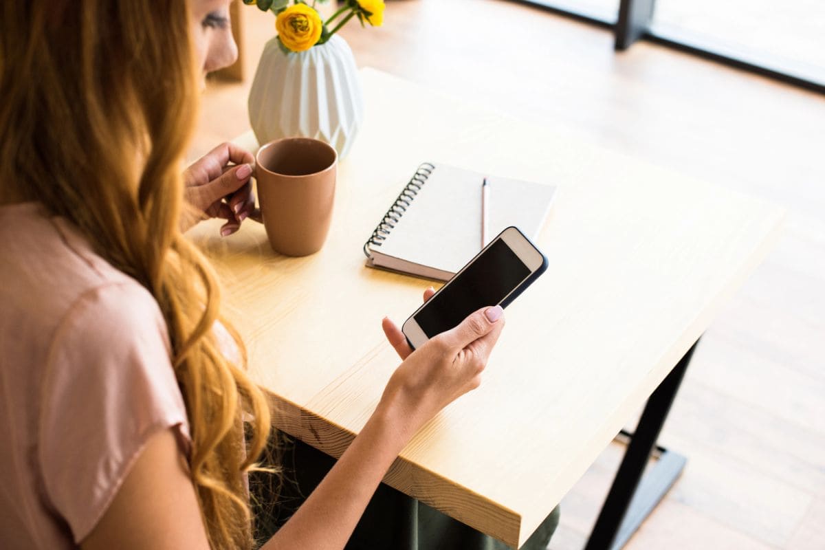 A woman sitting at a table, drinking coffee, and using a smartphone for blog post Qmee review