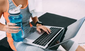 Woman wearing athletic gear and holding sports water bottle while working from home on a laptop.