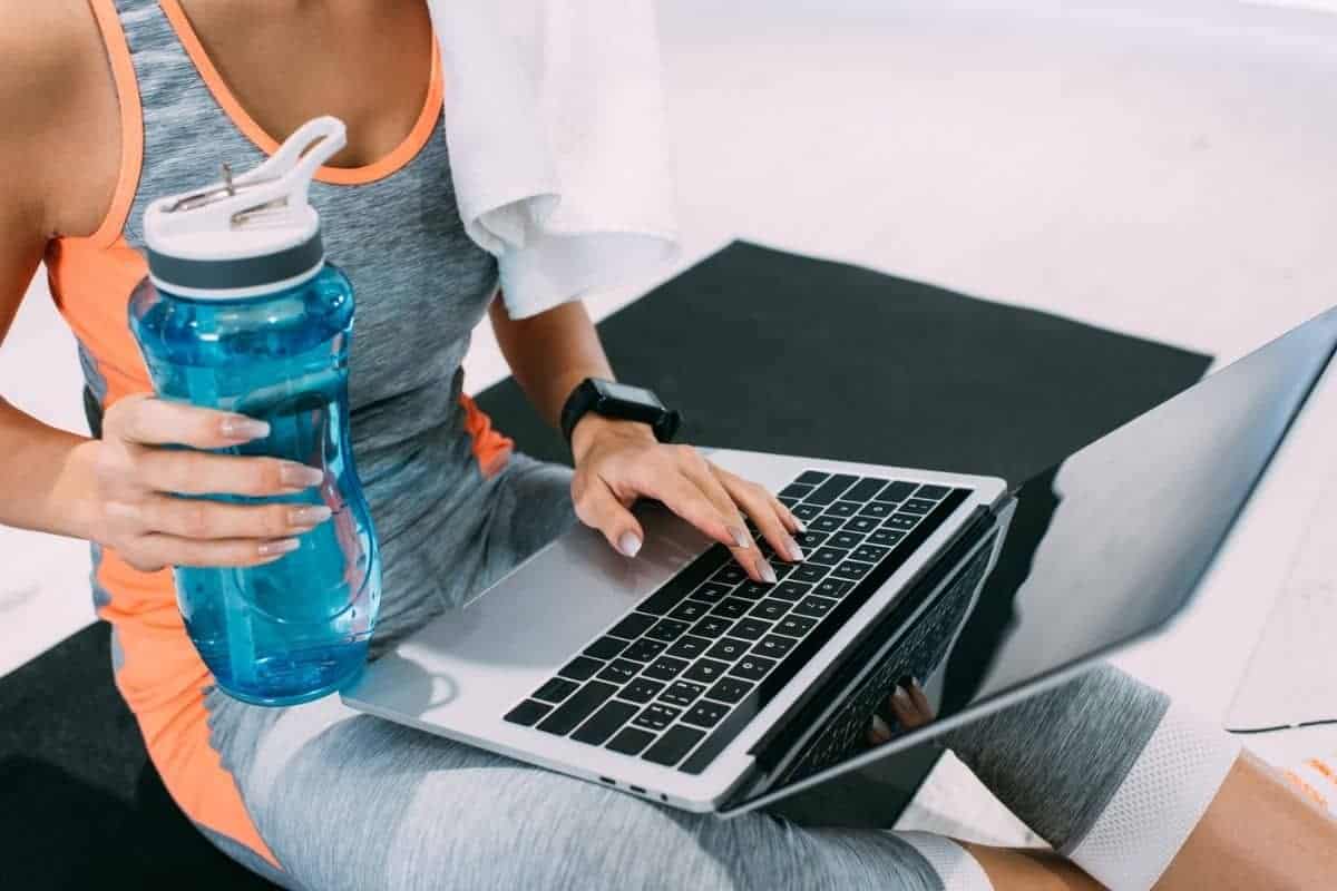 Woman wearing athletic gear and holding sports water bottle while working from home on a laptop.