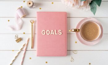 A notebook labeled, "Goals" sitting on a woman's desk, with coffee and flowers.