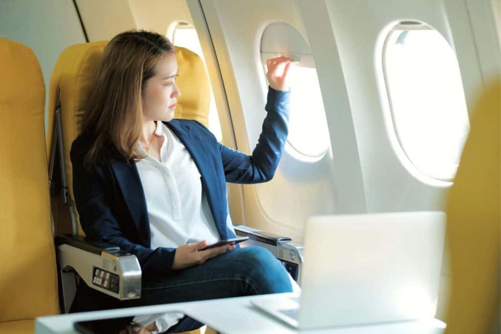 Businesswoman looking out of the window of an airplane, while working on a laptop.