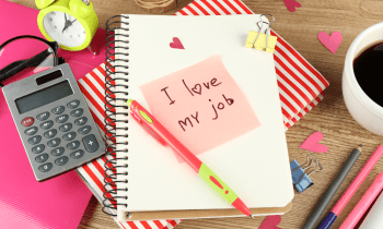 A stack of notebooks on a desk with some pens, a cup a coffee, and a note that says I love my job.