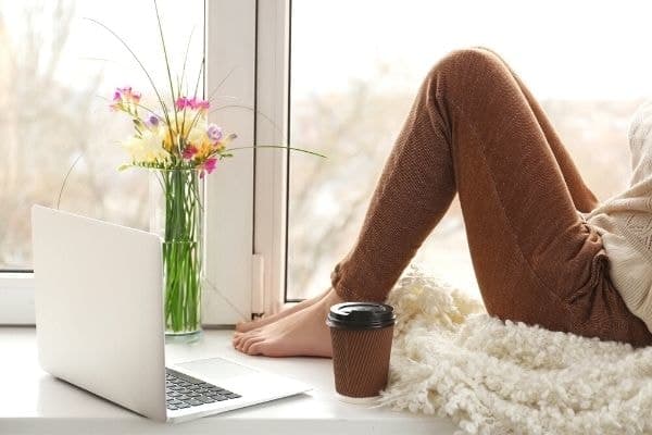 Woman sitting at home on window sill, working on laptop.