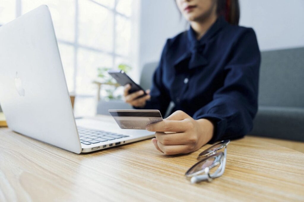 A woman is looking at the back of her credit card and using her laptop and phone to call her bank.