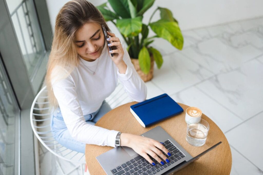 A young woman is working remotely using a laptop in a cafe and talking on the phone.
