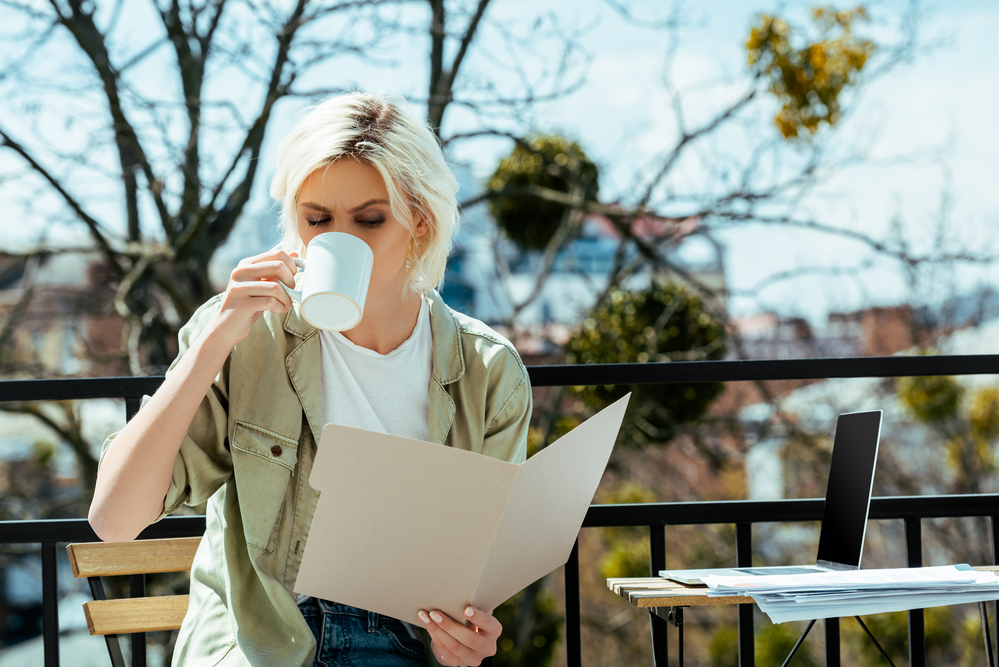 woman with blonde hair outdoors drinking coffee and reading a file folder with documents