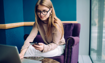 A young female wearing glasses looking at her phone and her laptop looking for free online courses for her work from home career