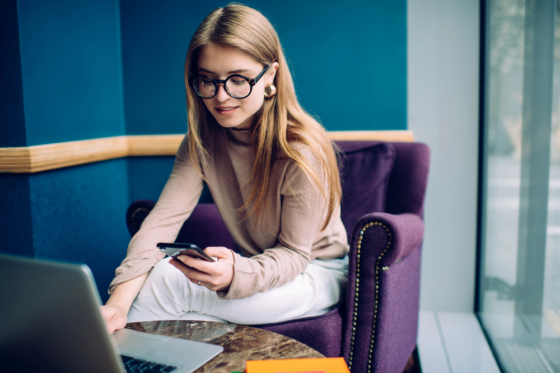 A young female wearing glasses looking at her phone and her laptop looking for free online courses for her work from home career