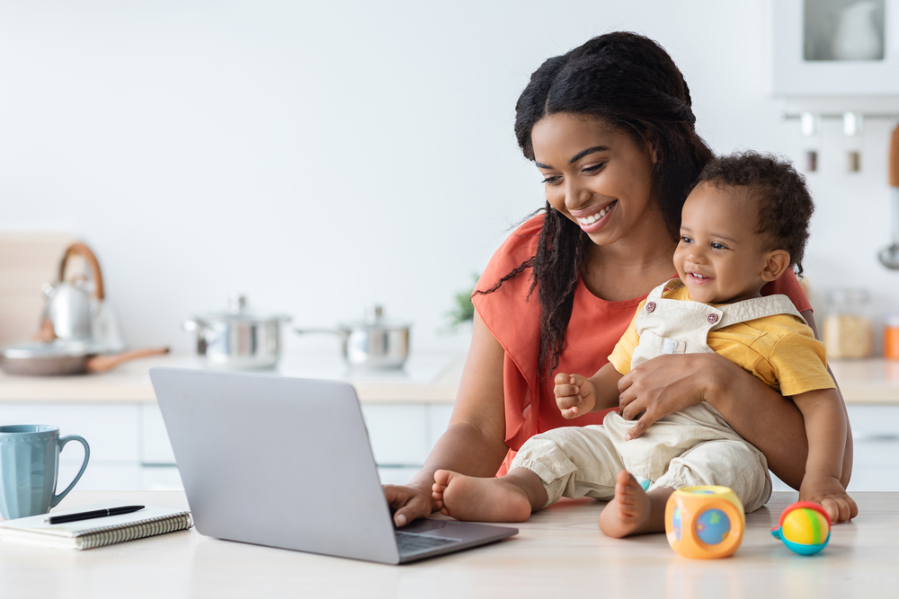 Young mom holding baby working on short tasks on her laptop
