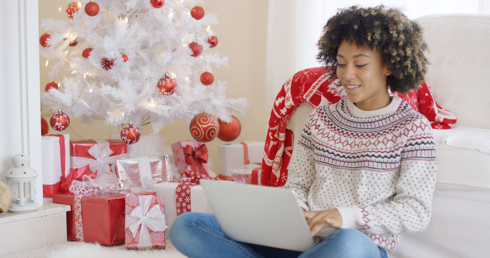 Young woman in a sweater working on laptop in front of Christmas tree